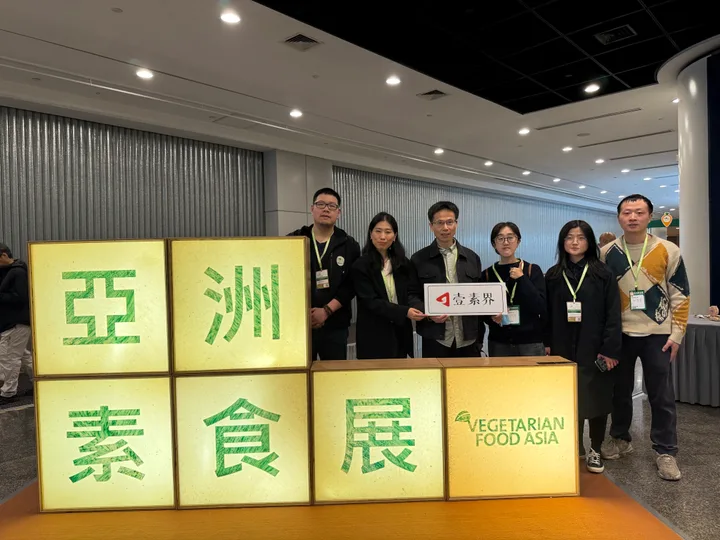 A group holding an allveg sign beside the Vegetarian Food Asia exhibition display.